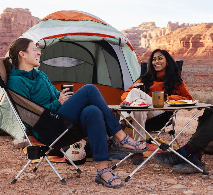 Two friends laugh and enjoy a meal together seated in camp chairs outside a tent in a desert landscape.